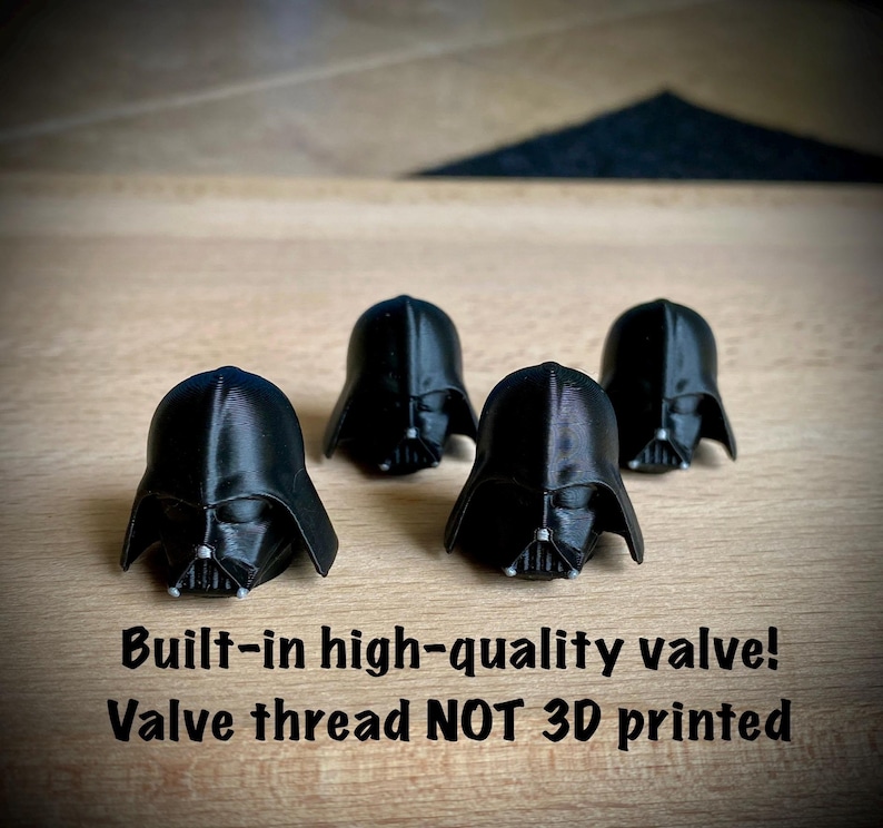 May include: Four black 3D printed helmet-shaped valve caps. The text below reads: Built-in high-quality valve! Valve thread NOT 3D printed. Each helmet features a smooth finish with a detailed face mask and a central silver-colored accent.