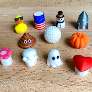 May include: A collection of eleven small decorative toppers including a yellow duck with sunglasses, an American flag cylinder, a snowman, a silver rocket, a brown poop emoji, a white golf ball, an orange pumpkin, a pink flower, a skull, a ghost, and a red heart.
