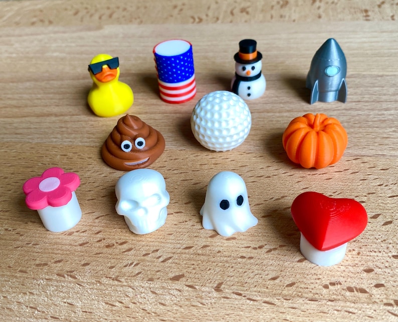 May include: A collection of eleven small decorative toppers including a yellow duck with sunglasses, an American flag cylinder, a snowman, a silver rocket, a brown poop emoji, a white golf ball, an orange pumpkin, a pink flower, a skull, a ghost, and a red heart.
