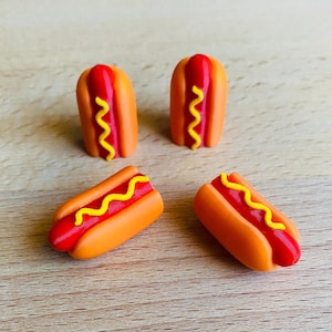 May include: Four miniature hot dogs with orange buns, red hot dogs, and yellow mustard. Two are upright, and two are lying down. The hot dogs are arranged on a light brown wooden surface.