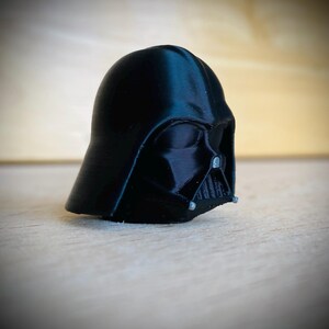 May include: A black 3D printed figurine of a helmeted character head. The helmet features a smooth, rounded dome, a triangular face mask with horizontal grill detailing, and small silver-colored accents on the lower sides of the mask.