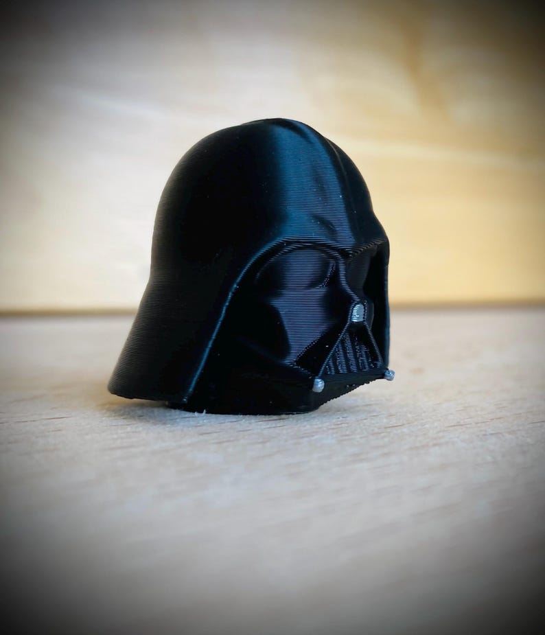 May include: A black 3D printed figurine of a helmeted character head. The helmet features a smooth, rounded dome, a triangular face mask with horizontal grill detailing, and small silver-colored accents on the lower sides of the mask.