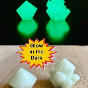 Könnte beinhalten: Zwei geometrische, kristallförmige Objekte, die im Dunkeln leuchten. Eines ist ein einzelner Diamant, das andere ein Cluster. Die Kristalle leuchten hellgrün. Das Bild zeigt die Kristalle auch in einem blassen, cremefarbenen Farbton.