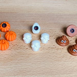 May include: A collection of small decorative charms including four orange pumpkins, four white skulls, and three brown smiling poop emojis with googly eyes. Each charm features a circular attachment point on the back for use as a shoe accessory.