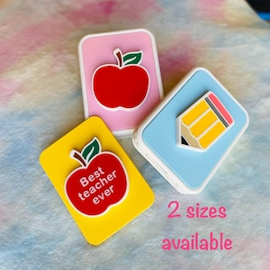May include: Three colorful rectangular charms with white borders. One is yellow with a red apple and the text "Best teacher ever." Another is pink with a red apple. The third is blue with a yellow pencil. The text "2 sizes available" is also visible.