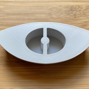 May include: White plastic eye-shaped mold with a small hole in the center. The mold is designed for making candles or soap.
