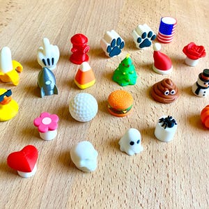 May include: A collection of twenty-two small, colorful, three-dimensional decorative toppers featuring various shapes including a banana, hand gesture, fire hydrant, paw print, American flag, rose, rocket, candy corn, Christmas tree, Santa hat, duck, golf ball, burger, poop emoji, snowman, flower, spider, heart, skull, ghost, and pumpkin.