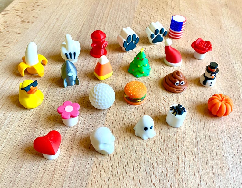 May include: A collection of twenty-two small, colorful, three-dimensional decorative toppers featuring various shapes including a banana, hand gesture, fire hydrant, paw print, American flag, rose, rocket, candy corn, Christmas tree, Santa hat, duck, golf ball, burger, poop emoji, snowman, flower, spider, heart, skull, ghost, and pumpkin.
