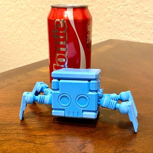 May include: A blue, 3D-printed robot spider toy with articulated legs, set in front of a red soda can. The robot has a blocky body with circular eye-like features. The soda can is red with white lettering.