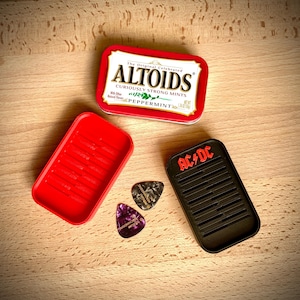 Könnte beinhalten: Eine rote Altoids-Minzschachtel, ein roter Kunststoffbehälter und ein schwarzer Gitarrenplektrumhalter mit dem AC/DC-Logo. Zwei Gitarrenplektren sind sichtbar, eines lila und eines schwarz-grau. Die Altoids-Dose ist geöffnet.