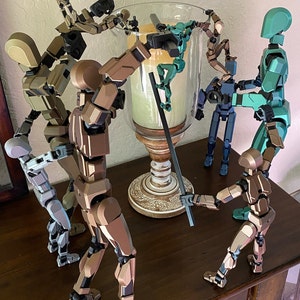 May include: A group of seven articulated figures made of plastic in various poses. The figures are painted in shades of brown, green, and blue. The figures are arranged around a glass candle holder with a white base. The figures are on a wooden table.