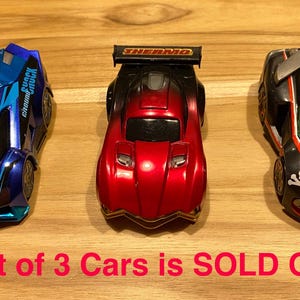 May include: Lot of 3 Cars is SOLD OUT. Three toy race cars, one blue, one red, and one black, with different designs.