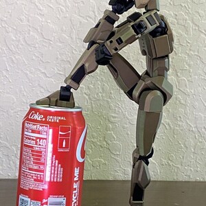 May include: A brown and tan articulated action figure with black joints is standing on a wooden surface. The figure is holding a can of Coca-Cola with the text "Coke Original Taste" and nutrition facts printed on the side. The can is red with white text and a white Coca-Cola logo.