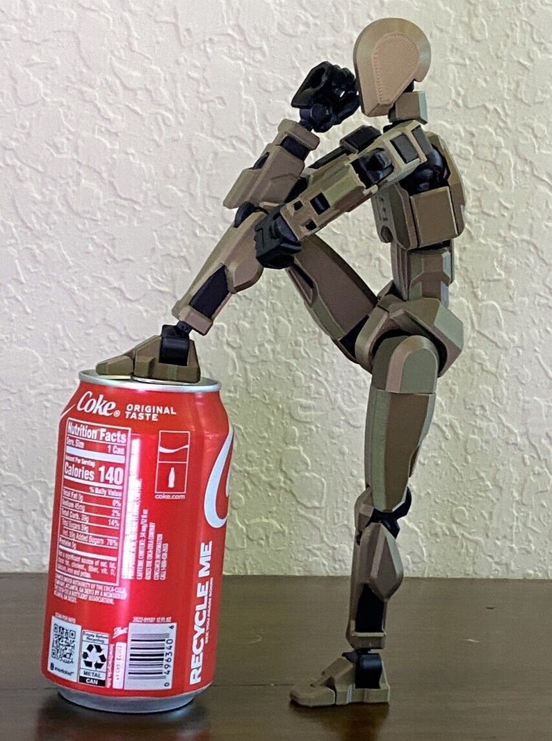 May include: A brown and tan articulated action figure with black joints is standing on a wooden surface. The figure is holding a can of Coca-Cola with the text "Coke Original Taste" and nutrition facts printed on the side. The can is red with white text and a white Coca-Cola logo.