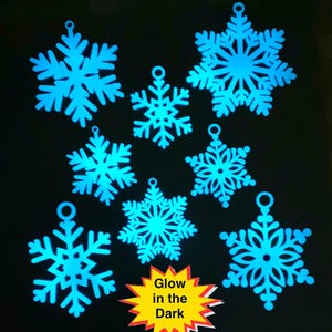 Może przedstawiać: Osiem niebieskich, świecących w ciemności ozdób w kształcie płatków śniegu. Płatki śniegu są wycięte z cienkiego materiału i mają delikatny wzór. Tekst "Glow in the Dark" jest nadrukowany na żółtej rozgwieżdżonej gwiazdce.