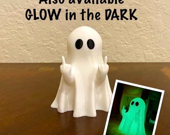 May include: A white ghost figurine with black oval eyes, holding up both middle fingers. The text above reads: Also available GLOW in the DARK. An inset image shows the ghost glowing green in the dark, confirming the glow-in-the-dark feature.