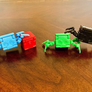 May include: Three small, articulated robot toys with different colored bodies. The robot on the left is blue, the middle robot is red, and the robot on the right is green. Each robot has six legs and a square body.