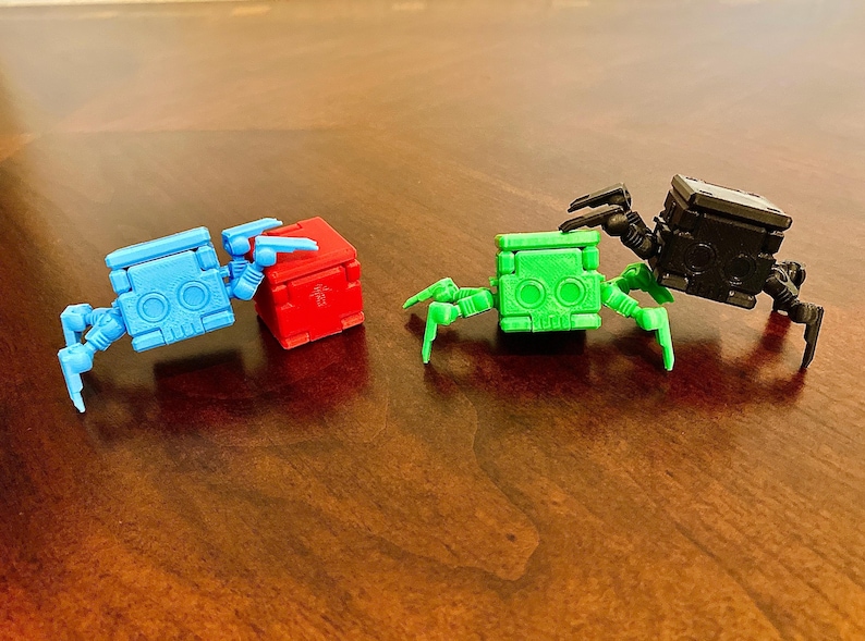 May include: Three small, articulated robot toys with different colored bodies. The robot on the left is blue, the middle robot is red, and the robot on the right is green. Each robot has six legs and a square body.