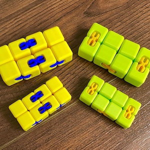 May include: Four small, colorful, cube-shaped fidget toys. Two are yellow with blue accents, and two are green with yellow accents.