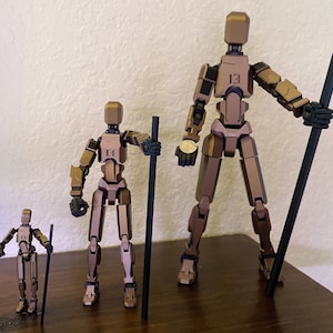 May include: Three articulated action figures made of brown plastic. The figures are standing on a wooden surface. The figures are holding black sticks. The tallest figure is holding a stick in its right hand and a coin in its left hand. The middle figure is holding a stick in its right hand. The smallest figure is holding a stick in its left hand.