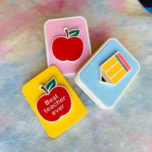 May include: Three colorful rectangular magnets. One is yellow with a red apple and the text "Best teacher ever". Another is pink with a red apple. The third is blue with a yellow pencil.