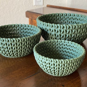 Puede incluir: Tres cestas tejidas verdes de diferentes tamaños. Las cestas están hechas de un material tejido que se asemeja a un patrón de punto. Las cestas están sobre una superficie de madera.