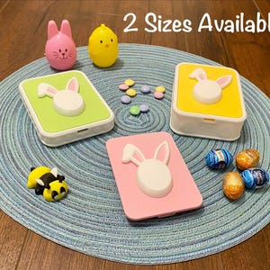 May include: Easter-themed decorative boxes in green, yellow, and pink, each featuring a white bunny design. The image also includes a pink bunny figurine, a yellow chick figurine, and colorful candies. Text reads "2 Sizes Available."