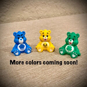 May include: Three small bear figurines in blue, yellow, and green. The blue bear has a cloud symbol, the yellow bear has a sun symbol, and the green bear has a clover symbol on their chests. The text below reads: More colors coming soon!