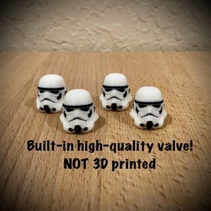 May include: Four white and black Stormtrooper helmet figurines are arranged on a wooden surface. The helmets have detailed black facial features. Text at the bottom reads "Built-in high-quality valve! NOT 3D printed."