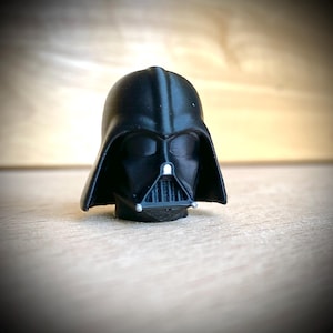 May include: A matte black 3D printed figurine of the Darth Vader helmet. The iconic headgear features a triangular nose section, a vertical grille mouth piece, and small silver-colored accents on the lower corners and center.
