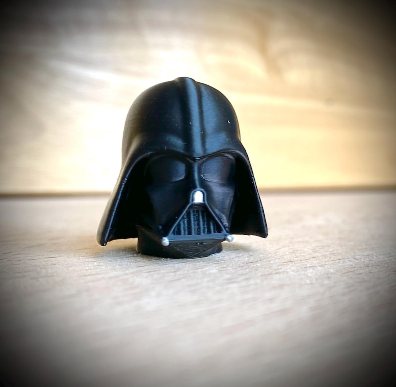 May include: A matte black 3D printed figurine of the Darth Vader helmet. The iconic headgear features a triangular nose section, a vertical grille mouth piece, and small silver-colored accents on the lower corners and center.