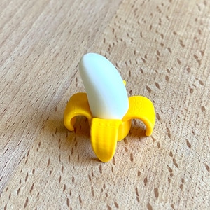 May include: A small, peeled banana ornament. The banana skin is a vibrant yellow, contrasting with the white banana flesh. It rests on a light brown wooden surface.