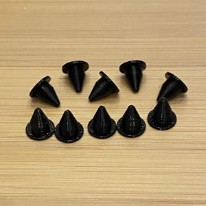 Puede incluir: Un conjunto de nueve tachuelas negras en forma de cono con base circular. Las tachuelas están dispuestas sobre una superficie de madera de color marrón claro. Las tachuelas tienen un aspecto texturizado y una punta.