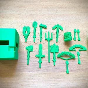 Puede incluir: Una colección de herramientas y accesorios de juguete de color verde brillante y pixelados, que incluyen un cubo, picos, espadas y una puerta. Los artículos están dispuestos sobre una superficie de madera de color marrón claro, lo que sugiere un tema de artesanía o construcción.