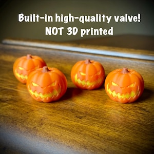 Op de afbeelding: Vier kleine oranje jack-o'-lantern pompoenen met geel uitgesneden gezichten. De tekst erboven luidt: Built-in high-quality valve! NOT 3D printed. Elke pompoen heeft een ronde vorm met verticale ribbels en een grillige, verlichte mond.