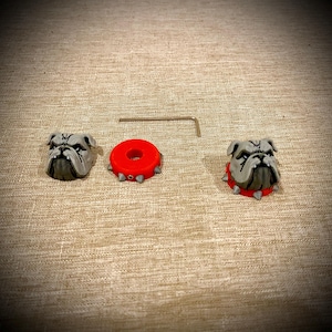 May include: Two gray bulldog head figurines with detailed facial features, one red circular collar with small silver spikes, and one L-shaped metal hex key tool. The bulldog heads are sculpted with visible wrinkles and dark markings.