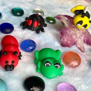 Puede incluir: Una colección de figuras coloridas impresas en 3D, que incluyen una oruga roja, una rana verde, una araña negra y una abeja amarilla. Las figuras están rodeadas de piedras de vidrio de varios colores sobre una superficie blanca y suave.