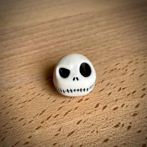 Puede incluir: Una figura blanca en forma de huevo con un ojo negro de forma ovalada, un ojo inclinado y una boca cosida. La figura es un personaje de Pesadilla antes de Navidad.