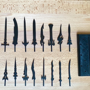 Puede incluir: Una colección de espadas negras en miniatura de varios diseños, dispuestas sobre una superficie de madera clara. Las espadas tienen diferentes formas de hoja y detalles de empuñadura. También hay un expositor verde oscuro con agujeros.