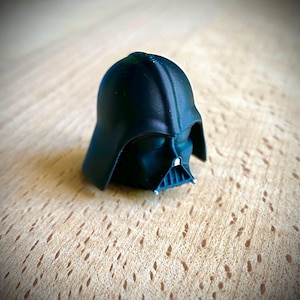 May include: A small, black, three-dimensional printed figurine of the Darth Vader helmet. The object features a smooth, rounded dome, a triangular face mask with a central grille, and visible horizontal layer lines from the printing process.