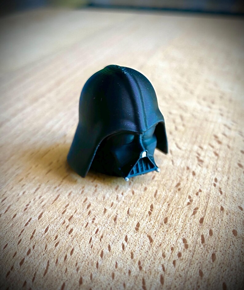 May include: A small, black, three-dimensional printed figurine of the Darth Vader helmet. The object features a smooth, rounded dome, a triangular face mask with a central grille, and visible horizontal layer lines from the printing process.
