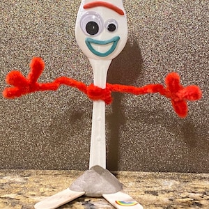 May include: A white plastic fork with a red and white striped headband, googly eyes, and a blue smile. The fork is attached to red pipe cleaner arms and legs, and is standing on a brown surface.