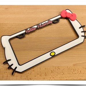 May include: White and black license plate frame with a Hello Kitty design. The frame features a pink bow, black whiskers, and the text "Hello Friends" in pink. A yellow circle is also present.
