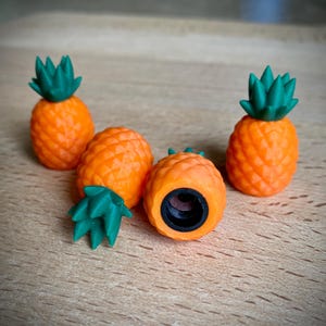 Op de afbeelding: Vier oranje ananasvormige ventieldopjes met groene bladeren. Eén dop is opengesneden en onthult de zwarte ventielsteel. De doppen zijn gerangschikt op een lichtbruin houten oppervlak.