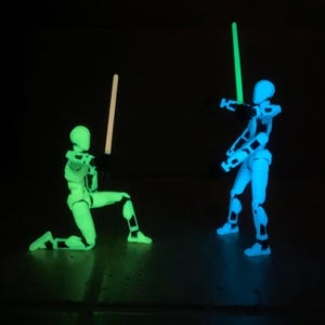 Puede incluir: Dos figuras de acción que brillan en la oscuridad sosteniendo espadas brillantes. Una figura está arrodillada y la otra está de pie. Las figuras están hechas de plástico blanco y tienen detalles negros. Las espadas son verdes y azules.