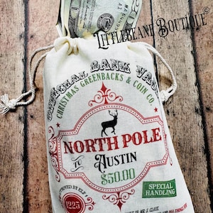May include: A cream-colored canvas drawstring bag featuring red, green, and black text and graphics. The design includes a reindeer silhouette, the text NORTH POLE TO AUSTIN, and $50.00. A twenty-dollar bill is visible protruding from the top opening.