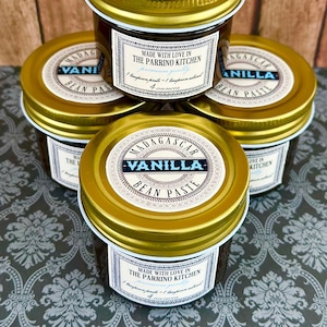 May include: Four glass jars with gold lids filled with dark brown vanilla bean paste. The jars are stacked on top of each other. Each jar has a label that says "Madagascar Vanilla Bean Paste" and "Made with love in The Parrino Kitchen".