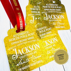 Set of high quality elegant gold glitter Santa Claus Gift tags  with holes punched and red Christmas ribbon on a white wood backdrop. The tags say" Authentic North Pole Post, Express Delivery Service, Special Delivery to: Jackson, Delivered By: Santa Claus, The maker of Holiday smiles