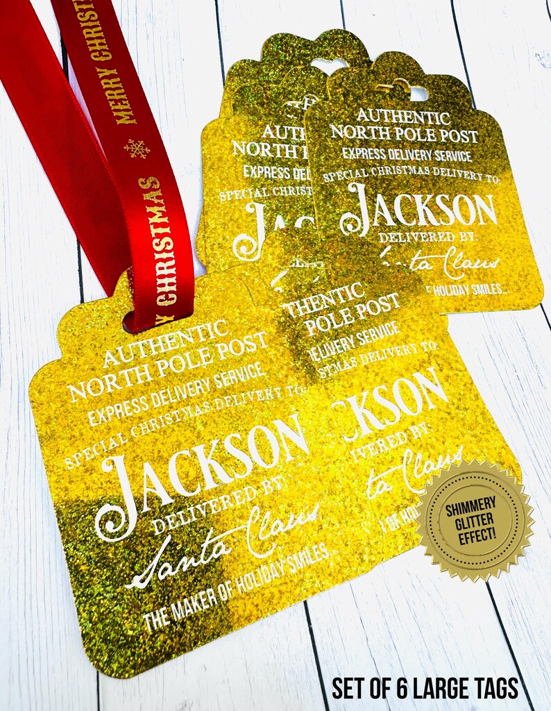 Set of high quality elegant gold glitter Santa Claus Gift tags  with holes punched and red Christmas ribbon on a white wood backdrop. The tags say" Authentic North Pole Post, Express Delivery Service, Special Delivery to: Jackson, Delivered By: Santa Claus, The maker of Holiday smiles