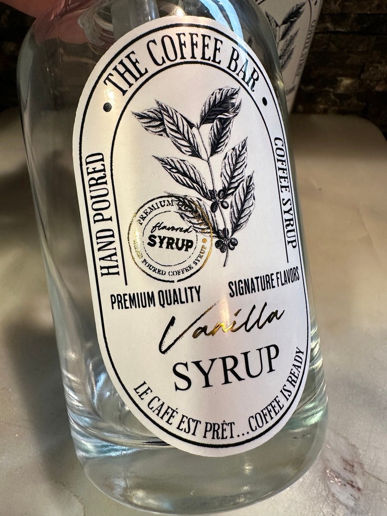 May include: A clear glass bottle with a white label that says "The Coffee Bar" and "Vanilla Syrup". The label also features a coffee bean branch illustration and the text "Hand Poured", "Premium Quality", "Signature Flavors", "Le Cafe Est Pret... Coffee Is Ready".